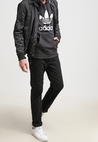 Svart nylonjacka över en svart huvtröja med vitt logotyp, tillsammans med svarta byxor och vita sneakers. Avslappnad look, minimala detaljer.