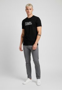 Czarny t-shirt z krótkim rękawem z szarym logo "KARL LAGERFELD", zestawiony z szarymi spodniami o dopasowanym fasonie i białymi trampkami na neutralnym tle.