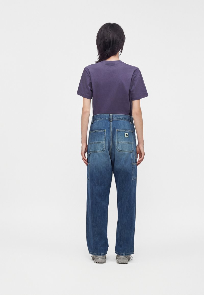 Person standing facing away wearing a purple short-sleeve shirt and loose-fitting blue jeans with back pockets and gray shoes.