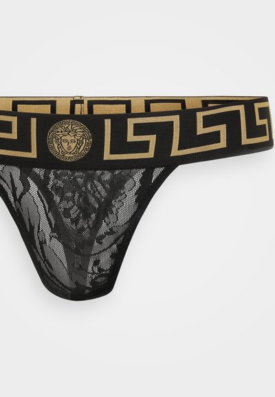 Zwarte kanten string met een brede gouden en zwart patroon tailleband met een Medusa-embleem en geometrische ontwerpen. Open gaastextuur.