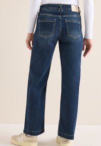 Unos jeans de mezclilla azul oscuro presentan un diseño de pierna recta, con una cintura de tiro bajo, cinco bolsillos y una textura de desvanecimiento sutil. Complementados con zapatillas.