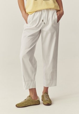Osoba ubrana w luźne białe spodnie o długości do połowy łydki z elastycznym pasem i sznurkami, zestawione z oliwkowo zielonymi trampkami na sznurowadła oraz żółtym topem.