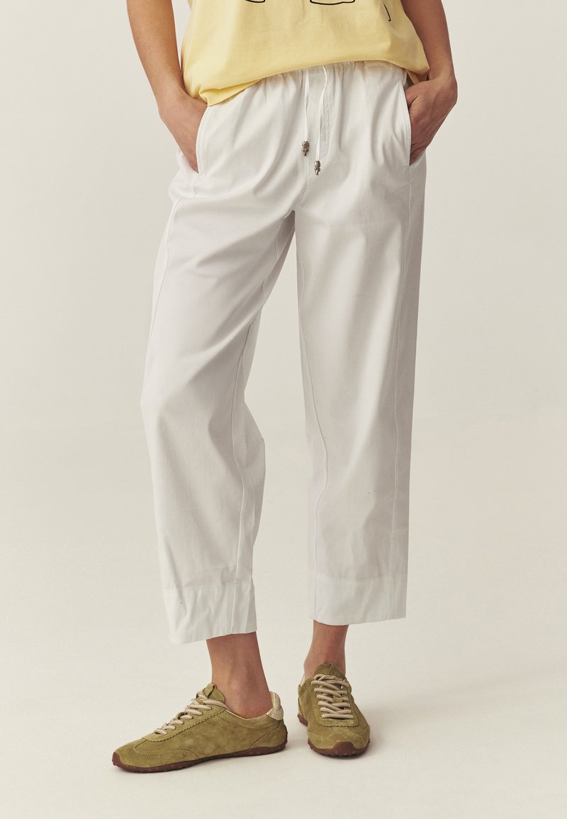 Osoba ubrana w luźne białe spodnie o długości do połowy łydki z elastycznym pasem i sznurkami, zestawione z oliwkowo zielonymi trampkami na sznurowadła oraz żółtym topem.