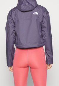 Chaqueta morada recortada con capucha y logo blanco en la espalda, combinada con leggings ajustados de color rosa. Tejido liso, líneas limpias y diseño minimalista.