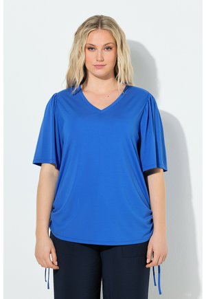 Kvinde med blondt, bølget hår iført en kongeblå V-hals skjorte med korte pufærmer og sorte bukser, stående mod en hvid baggrund.