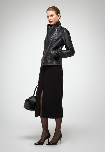 Giacca in pelle nera con zip asimmetrica, colletto alto e vestibilità strutturata. Abbinata a una gonna nera elegante con una fessura alta fino alla coscia.
