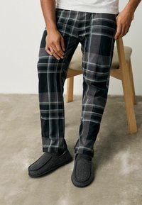 I pantaloni a quadri grigi presentano diverse tonalità di nero e grigio con una texture liscia. Le ciabatte nere in un tessuto testurizzato completano l'outfit.