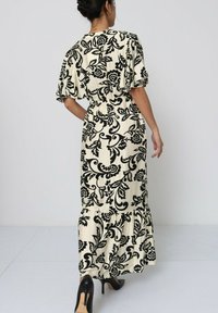 Robe en tissu crème avec un motif floral noir, des manches courtes et flottantes, une taille cintrée et une jupe évasée, associée à des talons hauts noirs.