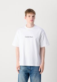 Balta medvilnės marškinėliai su trumpomis rankovėmis, apvaliu iškirpimu, su juodu "Calvin Klein" logotipu, centruotu ant krūtinės.