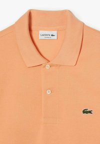 Polo tričko v broskyňovej farbe od značky Lacoste s klasickým strihom, s golierom, tromi gombíkmi a vyšitým logom krokodíla na hrudi.