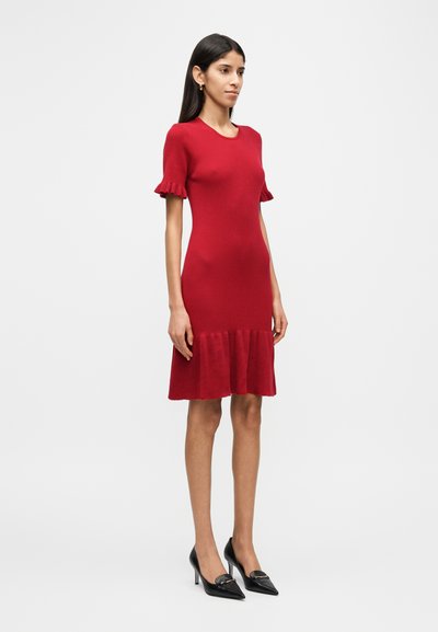 Κόκκινο πλεκτό φόρεμα με ριμπ ύφανση, κοντά μανίκια με βολάν, εφαρμοστό μπούστο και φαρδύ τελείωμα. Συνδυασμένο με μαύρες μυτερές γόβες.