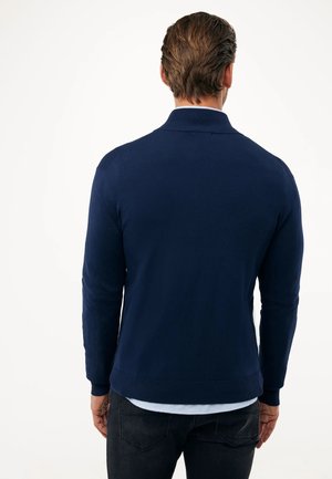 Marineblauwe gebreide trui met lange mouwen en een ribgebreide kraag. Effen textuur, aansluitend ontwerp, minimale details en een tailleband.