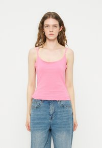 Γυναίκα με κυματιστά μαλλιά που φοράει χρυσά κρίκους στα αυτιά, ροζ τοπ με δαντέλα και τζιν με ψηλή μέση, στέκεται μπροστά σε απλό λευκό φόντο.