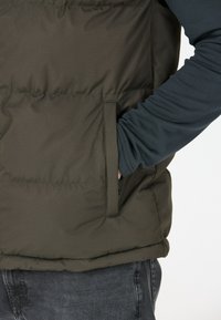 Dark green puffer vest with a smooth texture, featuring a side pocket and a high collar. Worn over a long-sleeve navy shirt.