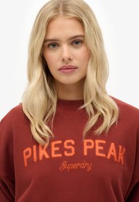 Sudadera granate de cuello redondo, con "PIKES PEAK" y "Superdry" bordados en naranja. Tejido suave con un corte relajado.