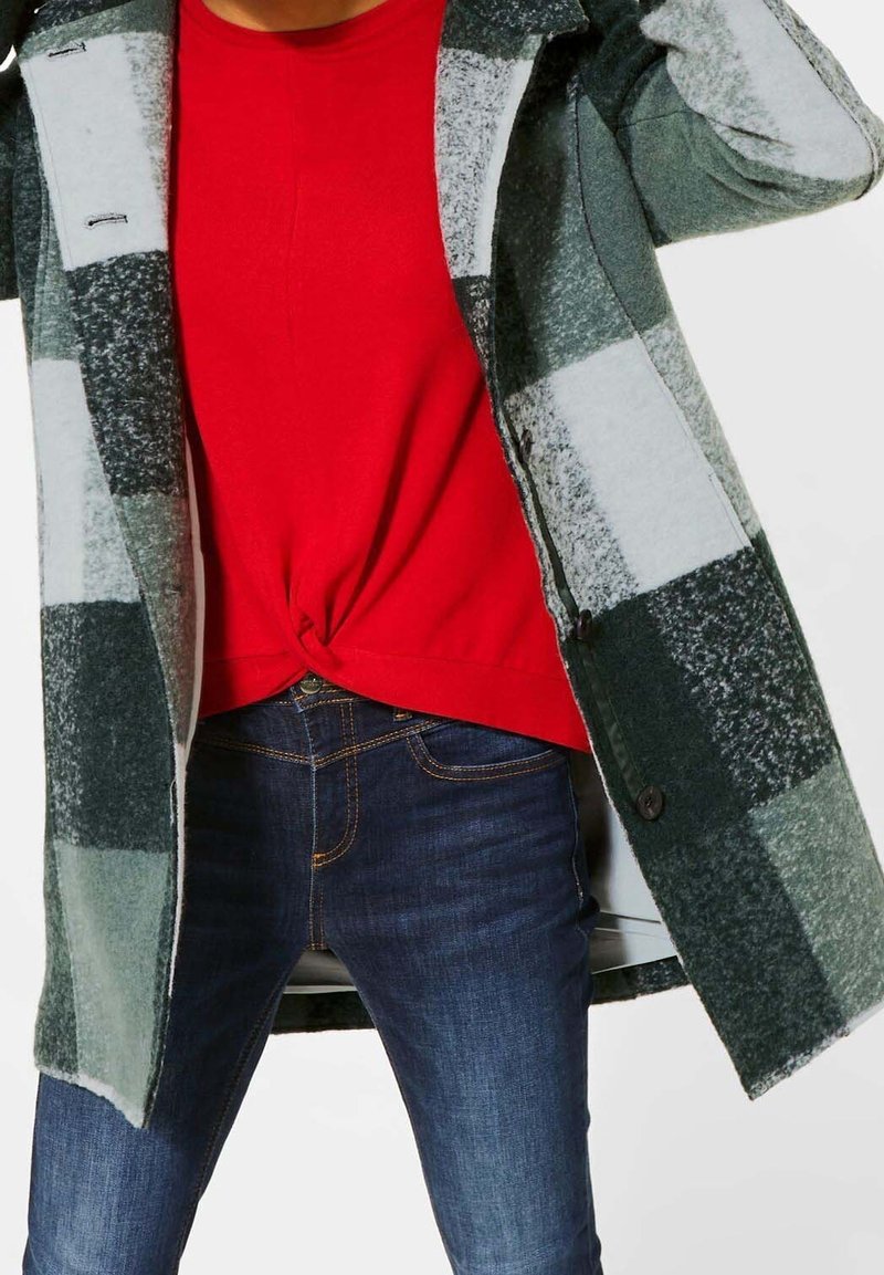 Grön och vit rutig ullrock med knäppningar, bärs över en röd skjorta, ihop med mörkblå jeans.