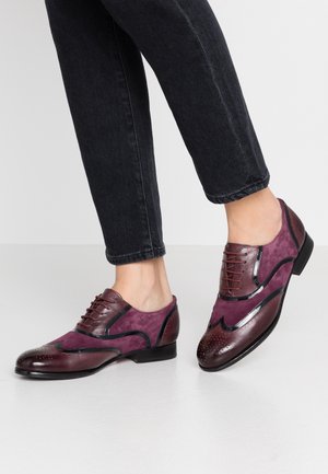 Person iført lilla og bordeaux læder- og ruskindsoxfordsko med sorte såler, kombineret med sorte cropped jeans.