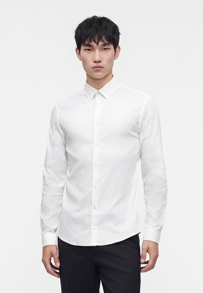 Young man wearing a fitted white button-up shirt and black pants, standing against a plain light background.