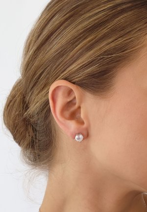 Runde Kristallstecker-Ohrringe mit einem silbernen Rahmen, die in einem durchstochenen Ohr platziert sind und eine klare, facettierte Oberfläche zeigen.