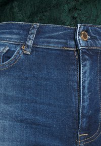 Limpar calças de ganga azul com uma cintura alta, apresentando um botão de metal, detalhes em costura dourada e uma textura suave e ligeiramente desgastada.