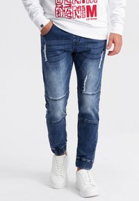 Sinimuotoiset siniset farkut, joissa on kulutettu ulkoasu, polvikohdat ja vaalea pesu, yhdistettynä valkoisiin tennareihin.