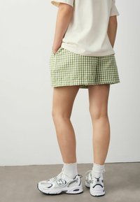 Grønne og hvide gingham shorts med sidelommer, parret med hvide New Balance sneakers og ankelstrømper. Glat bomuldsstruktur.