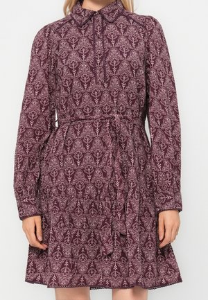 Frau trägt ein langärmeliges, dunkelrotes Kleid mit Blumenmuster, hohem Kragen und Taillengürtel, steht vor einem schlichten weißen Hintergrund.