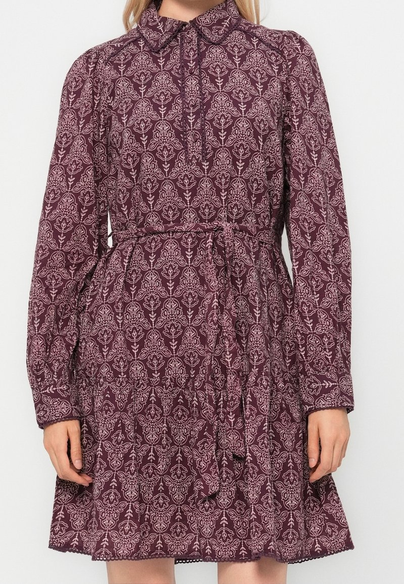 Femme portant une robe longue à manches longues, bordeaux avec un motif floral, col montant et ceinture à la taille, debout devant un fond blanc uni.