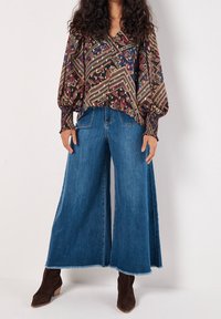 Blouse à motifs avec manches bouffantes, associée à un jean denim bleu à jambes larges avec ourlets effilochés et bottines marron à talons.