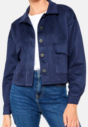 Marineblaue, kurz geschnittene Jacke mit Kragen, vorderer Knopfleiste, zwei Brusttaschen und weicher Textur. Kombiniert mit blauen Denim-Jeans.