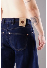 Blaue Jeans aus Denim mit kupfernen Nieten, Gesäßtaschen und einem beigen Lederlabel am Bund mit Markenlogo und Text.