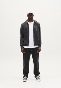 Gray velour zip-up hoodie with kangaroo pockets, paired with a white t-shirt and black loose-fit pants. Tan baseball cap and striped shoes.