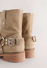Beige Wildleder-Stiefeletten mit Schnallenriemen, flachem Holzabsatz und glatter Textur. Verfügt über Doppelnaht und silberne Metallakzente.