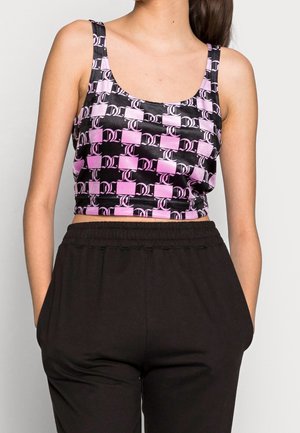 Vrouw draagt een roze-zwart patroon crop top en losse zwarte broek met handen in de zakken tegen een effen achtergrond.