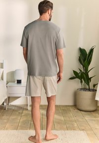 Szary T-shirt z krótkim rękawem w połączeniu z pasiastymi beżowymi szortami. Miejsce w pokoju z drewnianą podłogą, doniczką oraz lampą w tle.