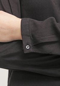 Sort skjorte med knaplukning i detaljeret tekstil. Har en manchet med en enkelt rund knap og en glat, ren finish.