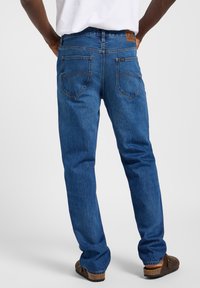 Vaqueros de mezclilla azul con corte recto, con bolsillos traseros y un parche de cuero en la cintura. La tela tiene un lavado claro y una textura suave.