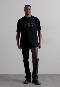 T-shirt noir avec plusieurs logos, coupe décontractée. Associée à un pantalon en cuir noir élégant et des chaussures à lacets noires. Fond uni.