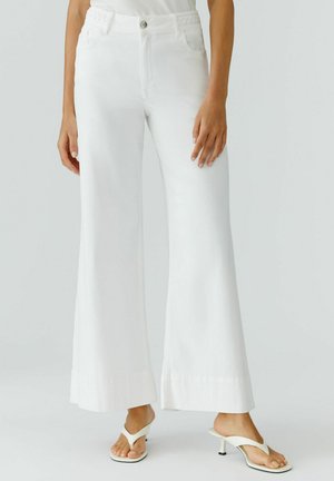 Vrouw draagt witte, hoge taille, brede broekspijpen en witte sandalen met hakken, staand tegen een effen lichte achtergrond.