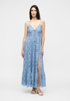 Γυναίκα που φοράει ένα αμάνικο ανοιχτό μπλε δαντελένιο φόρεμα με V-λαιμόκοψη, δέσιμο στο μπροστινό μέρος, ψηλή σχισμή και βολάν στο τελείωμα, συνδυασμένο με λευκά σανδάλια με λουράκια.