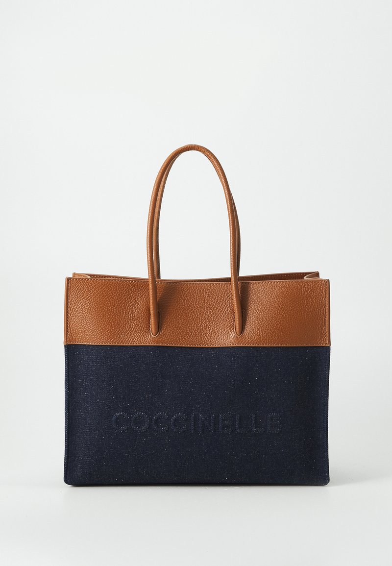 Vegetable-tanned leather and denim tote bag with brown top, navy body, embossed logo on the front, and two sturdy handles.