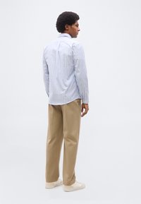 Άνδρας με αφροκέντικο χτένισμα, φορώντας μπλε και λευκό ριγέ πουκάμισο, μπεζ παντελόνι και λευκά αθλητικά παπούτσια, στέκεται μπροστά σε απλό φόντο.