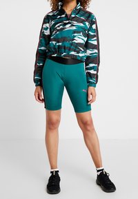 Kamouflage-mönstrad jacka med dragkedja i svart, teal och vitt; matchad med teal atletiska shorts. Båda har resårmidja och varumärkesdetaljer.