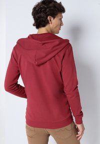 Sweat à capuche rouge avec un cordon de serrage et des poignets côtelés, fabriqué à partir d'un tissu doux, avec une texture lisse et une coupe décontractée.