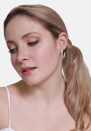 Goldene Creolen mit eingelassenen klaren Kristallen, die eine strukturierte Oberfläche und ein elegantes Design aufweisen. Das Model hat lange, hellbraune Haare, die zu einem Pferdeschwanz gebunden sind.