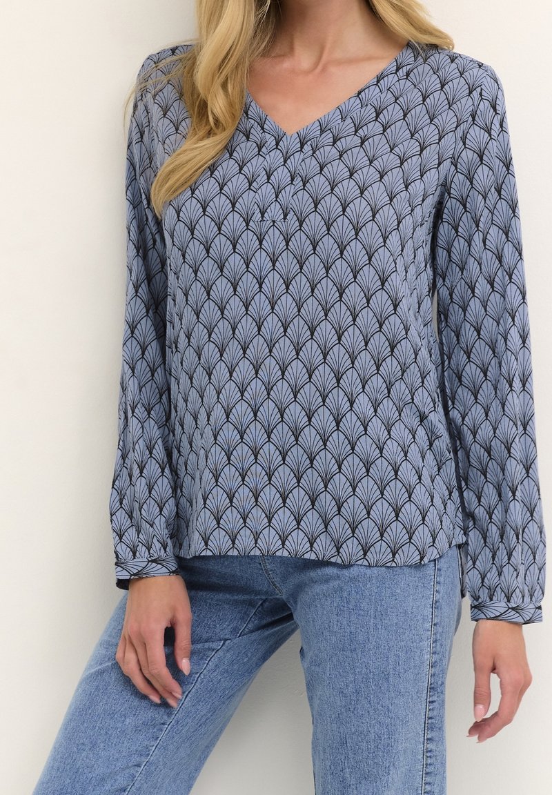 Blouse en V en tissu bleu avec un motif de feuilles noires. Manches longues avec poignets boutonnés. Associée à un jean en denim clair.