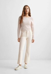 Maglione a coste rosa chiaro con scollatura a girocollo, abbinato a jeans cremosi a trapezio. Il modello indossa sneakers di colore chiaro. Texture morbide, stile casual.