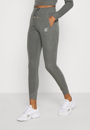 Donna con joggers grigi aderenti con logo bianco sulla coscia e sneaker bianche, una mano in tasca, in piedi contro uno sfondo semplice.