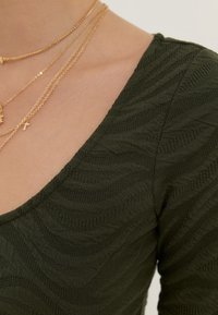 Tessuto verde scuro con una trama a onde, caratterizzato da un ampio scollo. Collane d'oro a strati accentuano l'area della clavicola.