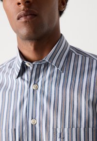 Csíkos ing kék és barna függőleges csíkokkal, hegyes gallérral és két elülső zsebbel, melyeken logó részlet található.